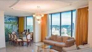 A modern living room with a glass coffee table, beige sofa, and dining area with chairs. Large windows offer a city view. - Hotel Golden Park Internacional Foz & Convenções By Nacional Inn (Foz do Iguaçu)