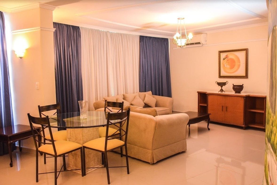 A living room with a glass-top dining table, beige sofa, and dark curtains.