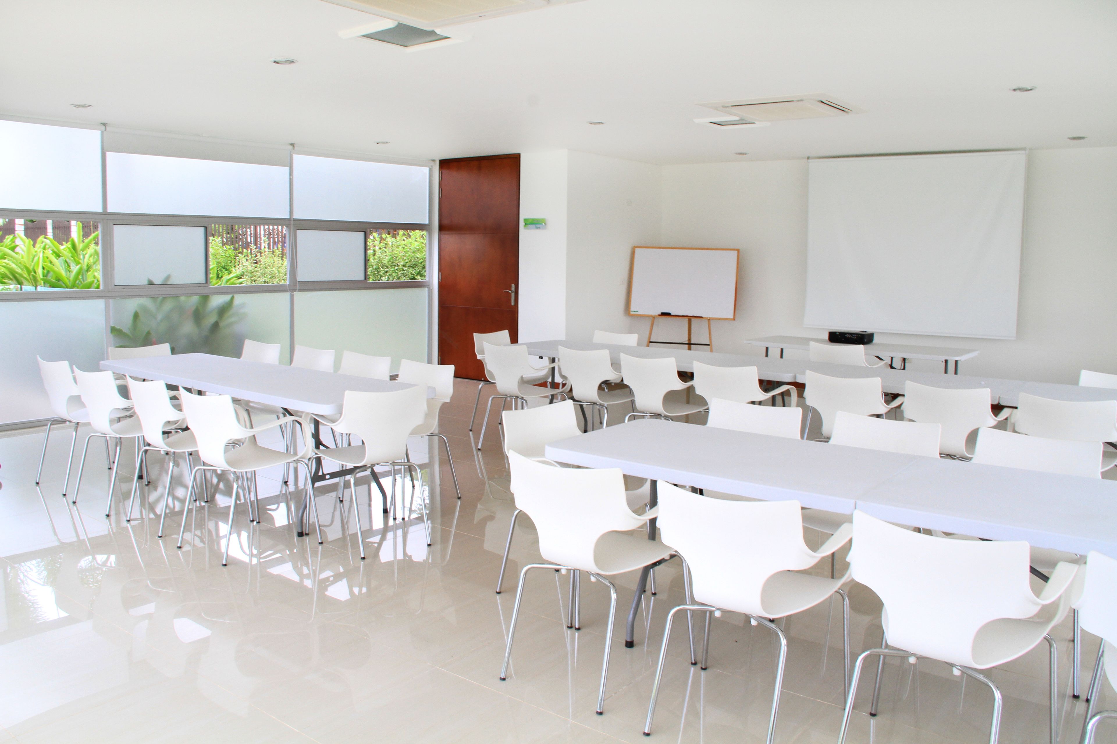 Uma sala de conferências com mesas e cadeiras brancas, uma lousa branca e um projetor.