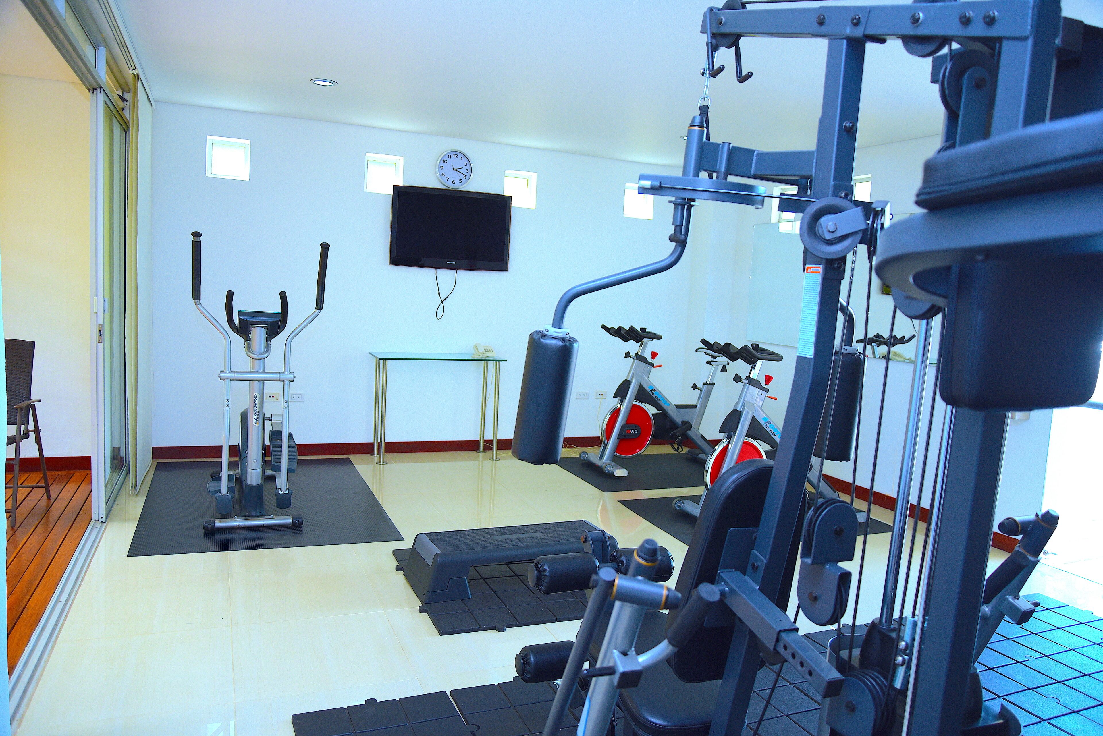 Uma academia com equipamentos de exercícios, uma televisão na parede e um relógio.