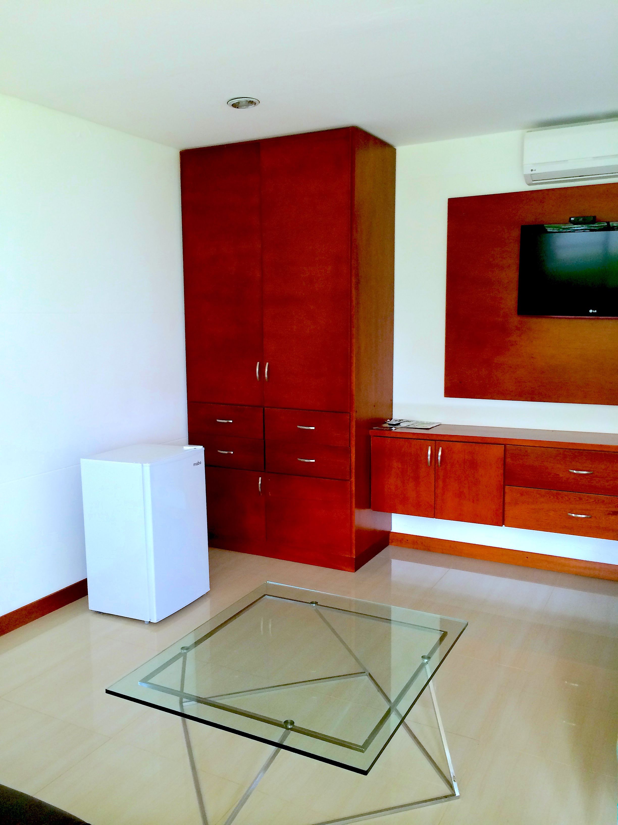 A compact kitchen with a white refrigerator, a glass-top table, red cabinets, and a mounted TV.