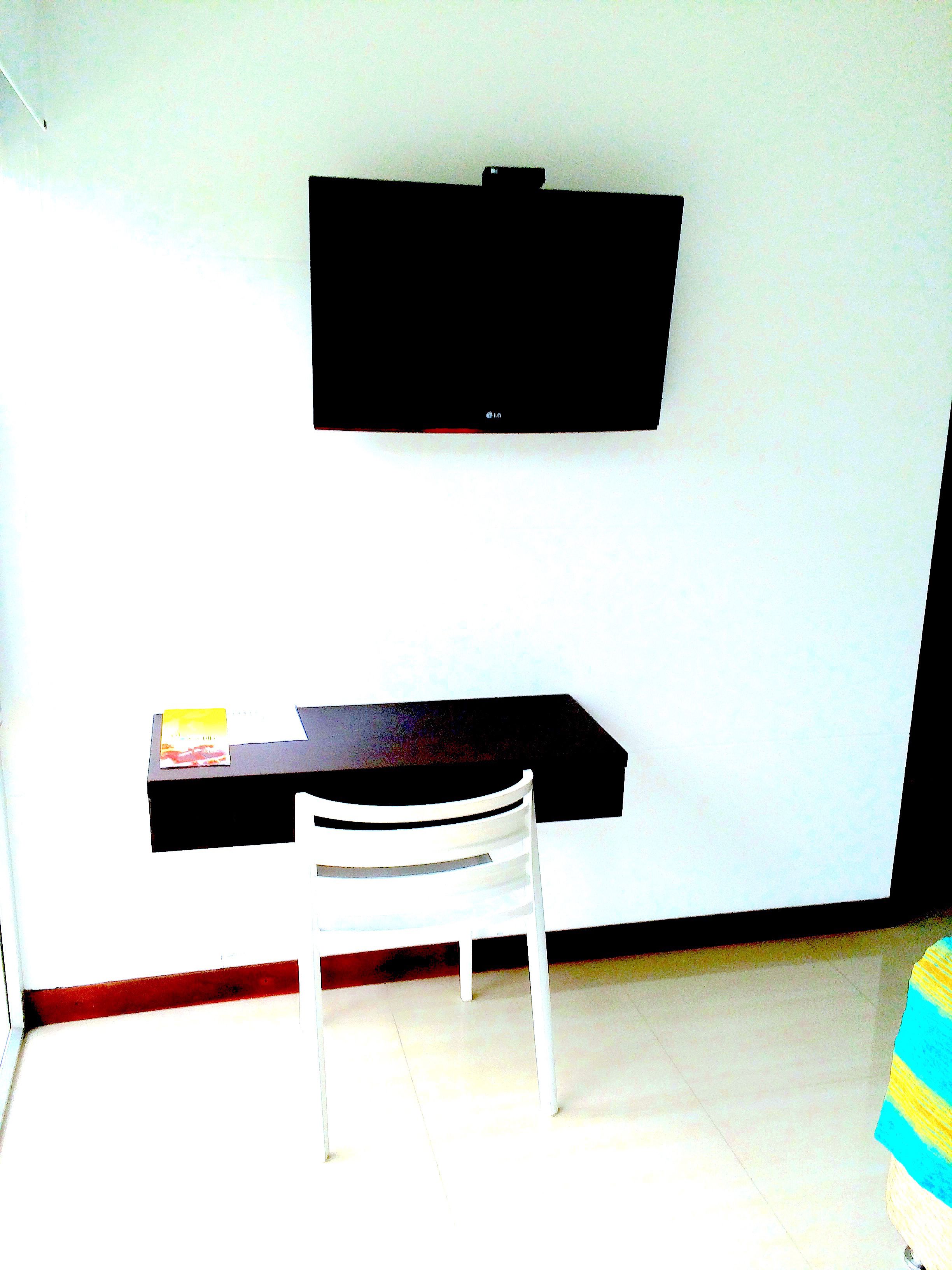 A room with a flat-screen TV mounted on the wall, a desk with a chair, and a small table with a book.