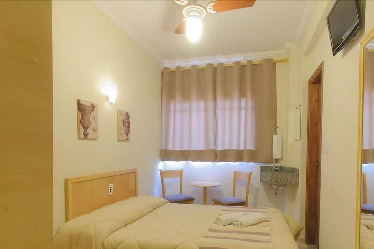 Ein Hotelzimmer mit Bett, kleinem Tisch, Stühlen, Deckenventilator, Fernseher und einem Fenster mit Vorhängen.