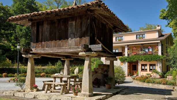 A rustic wooden structure with a stone bench and potted plants, situated in a garden area with a building in the background. - Casa Rural Santu Colás (Cangas de Onis)