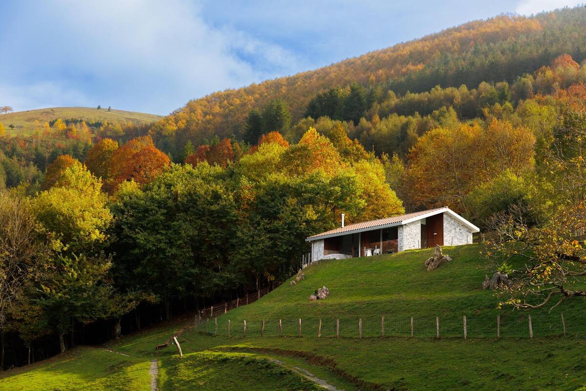 Une maison en pierre avec un toit en pente, située sur une colline verdoyante et entourée d’arbres aux couleurs automnales.