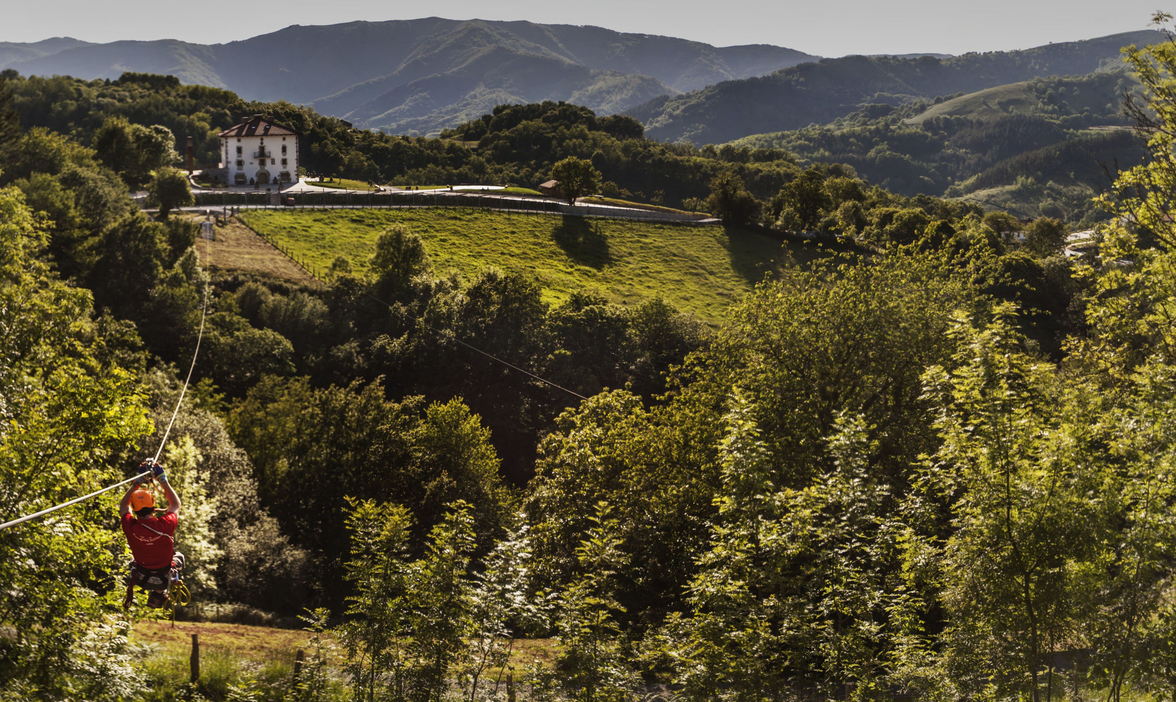 A scenic view of a lush green valley with a white building in the distance, surrounded by trees and hills.