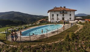 Une piscine entourée d’une clôture, avec un bâtiment en arrière-plan et des montagnes au loin.