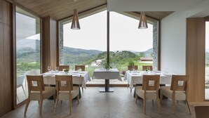 A dining area with a view of the countryside, featuring wooden tables and chairs, large windows, and pendant lighting.
