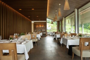 Un restaurant avec des boiseries, des nappes blanches et de grandes fenêtres donnant sur l’extérieur.