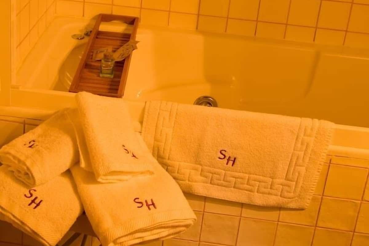 Kylpyhuone, jossa on kylpyamme, puinen tarjotinalusta ja kolme pyyhettä, joihin on kirjottu kirjaimet »SH».