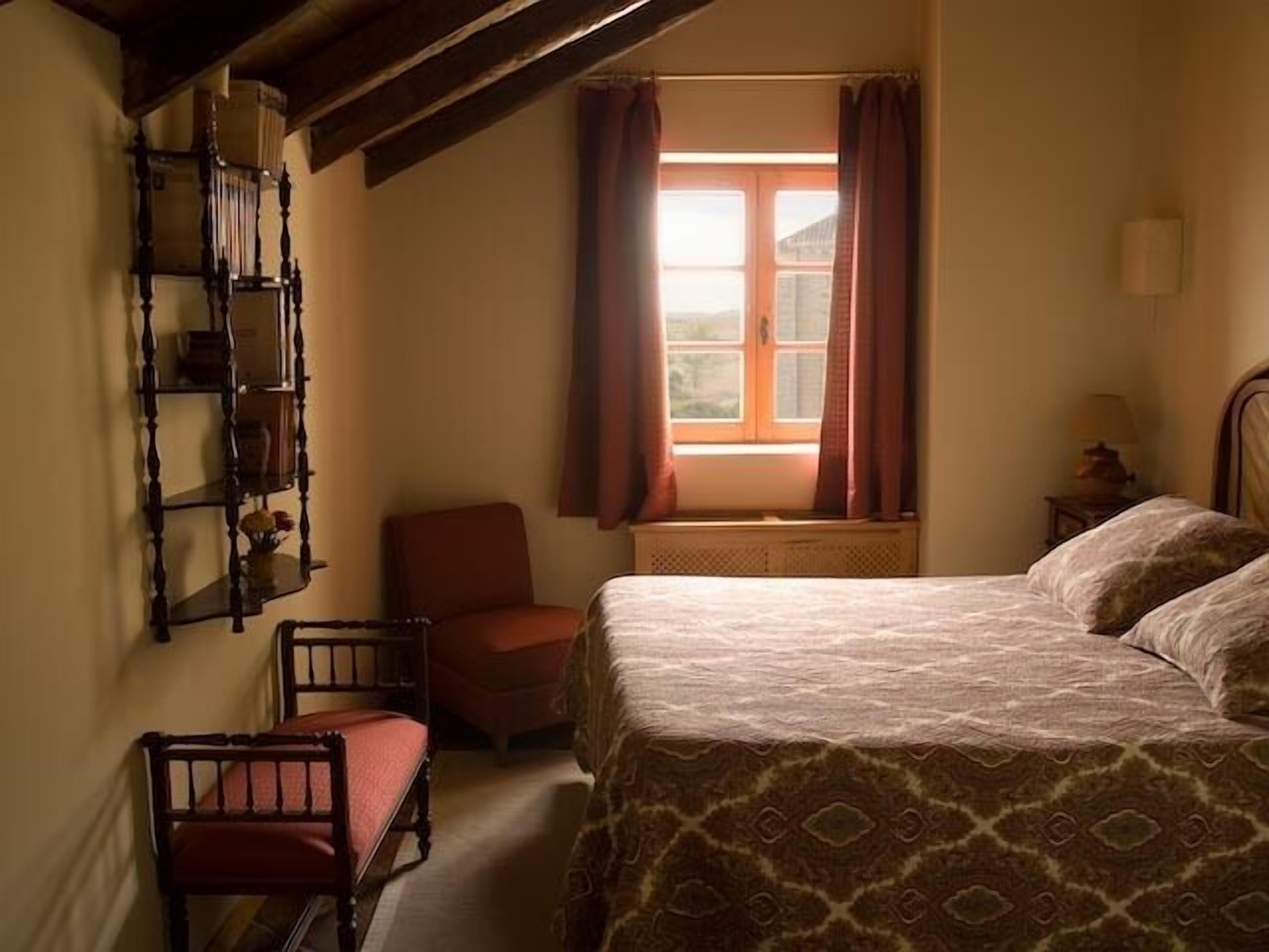 Une chambre à coucher avec un lit, une chaise, une fenêtre avec des rideaux, une étagère avec des livres et une lampe.