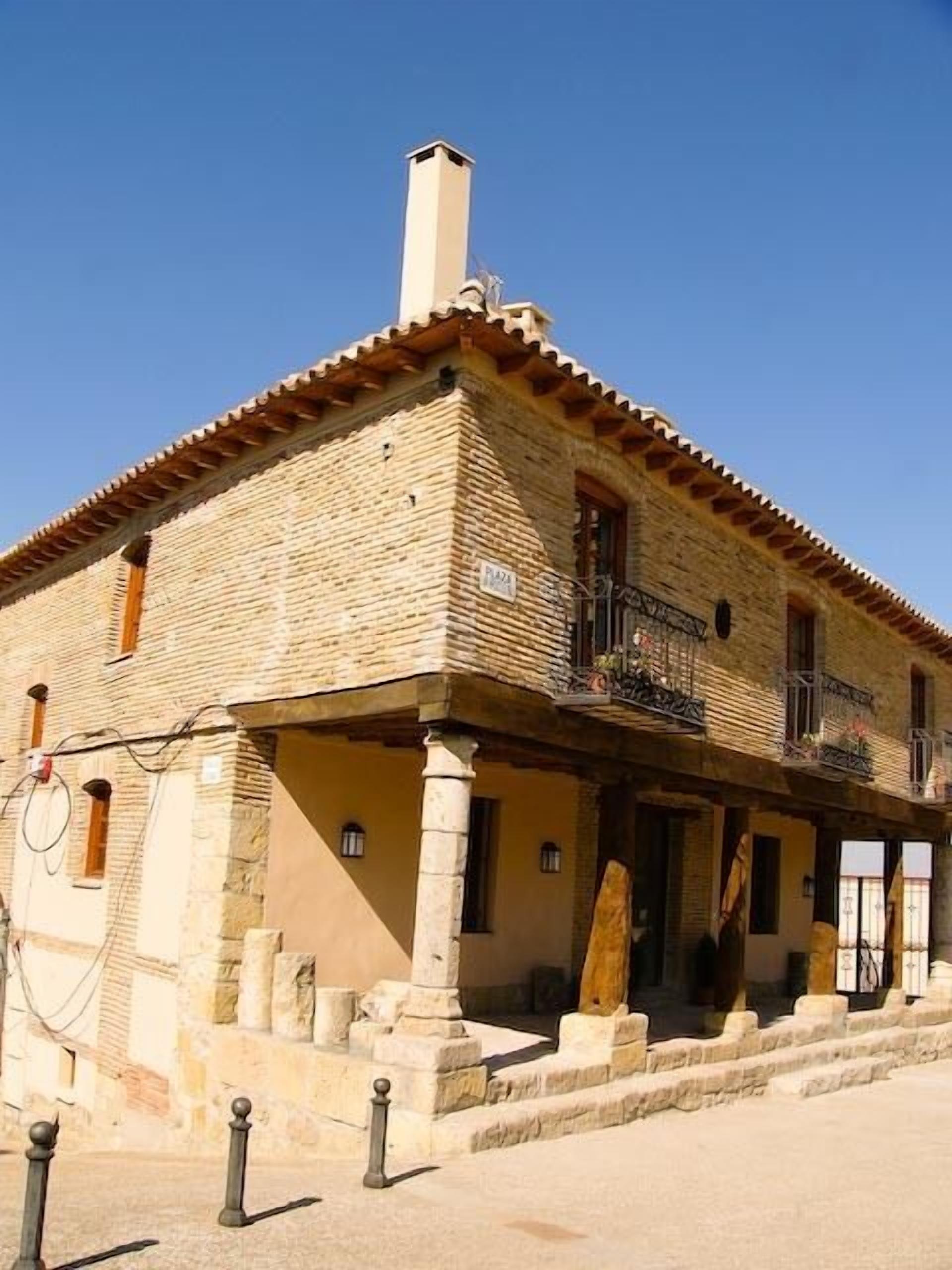 A traditional stone building with a tiled roof, a balcony with a wrought iron railing, and a sign indicating 'Plaza'.
