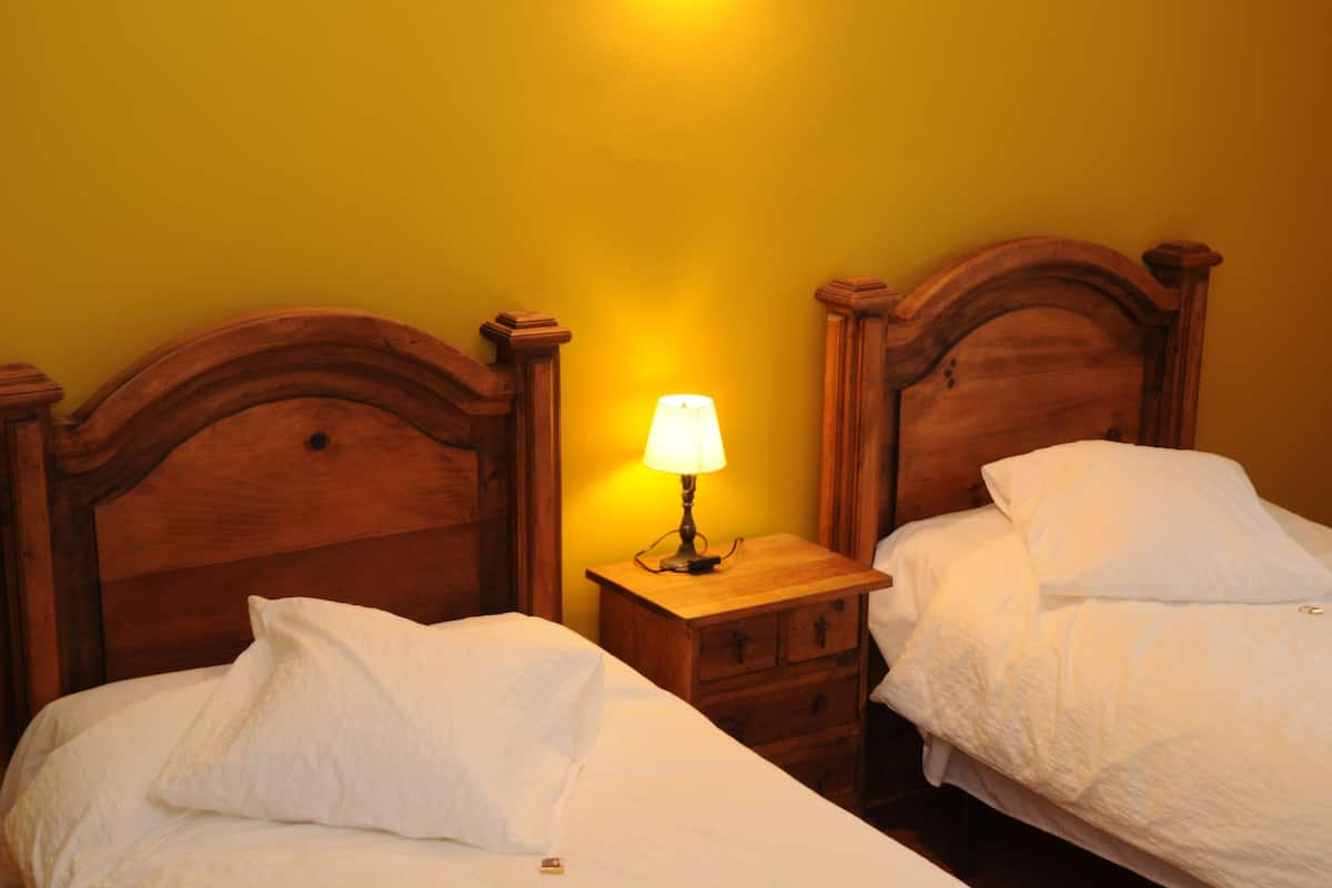 Quarto com duas camas de madeira, cada uma com roupa de cama branca, e uma mesa de cabeceira com abajur entre elas.