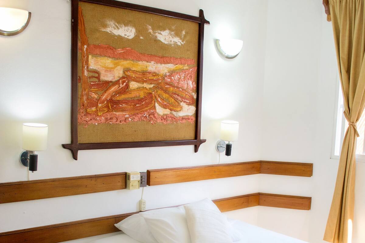 Una habitación de hotel con cama, mesita de noche, un cuadro en la pared y dos lámparas fijadas a la pared.
