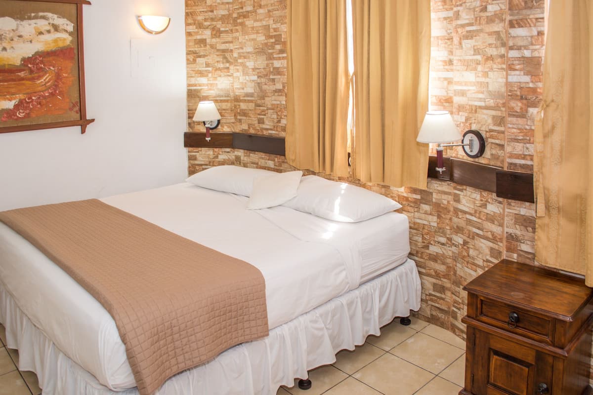 Una cama bien hecha con una manta marrón, dos lámparas de noche, un mesita de noche de madera y un reloj de pared.