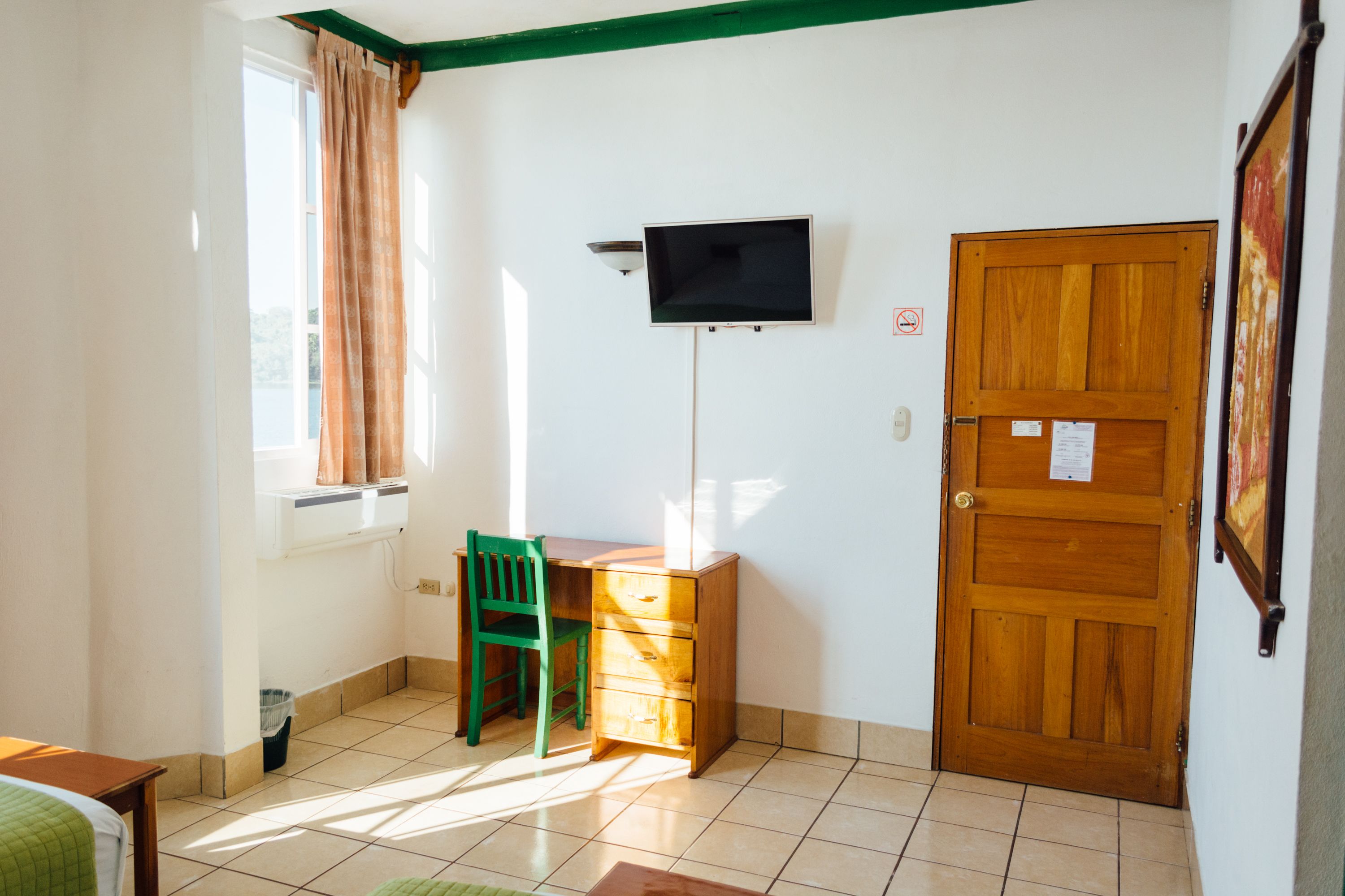 A small room with a wooden desk, a green chair, a television mounted on the wall, and a wooden door.