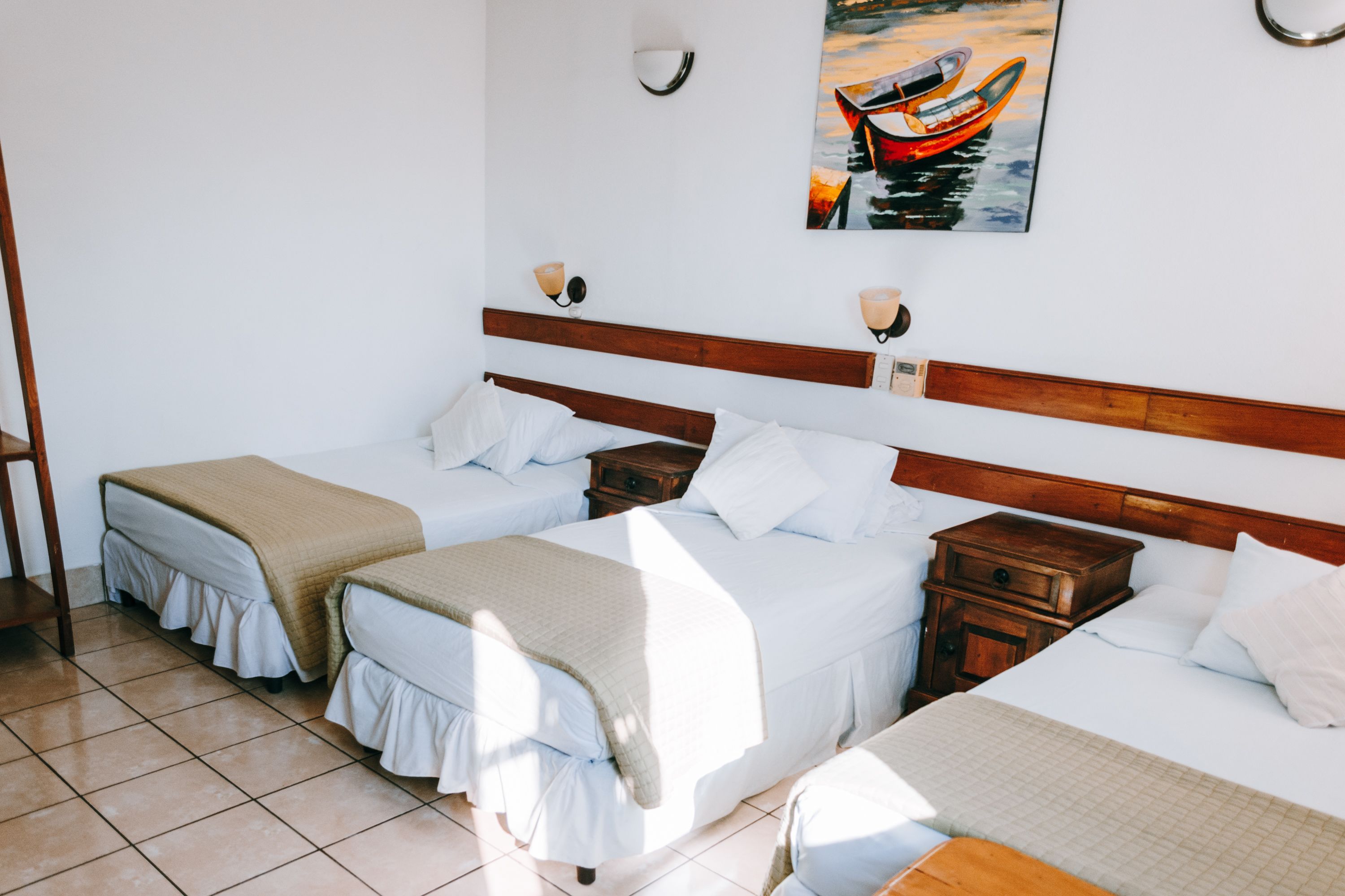A hotel room with two single beds, a wooden headboard, a nightstand, a small table, and a wall-mounted painting of a boat.