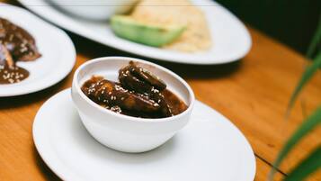 Un cuenco blanco con comida sobre un plato blanco, con un fondo desenfocado de una mesa y otro plato con un elemento verde.