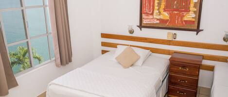 A hotel room with a bed, a wooden dresser, a nightstand, a window with a view of the sea, and a painting on the wall.