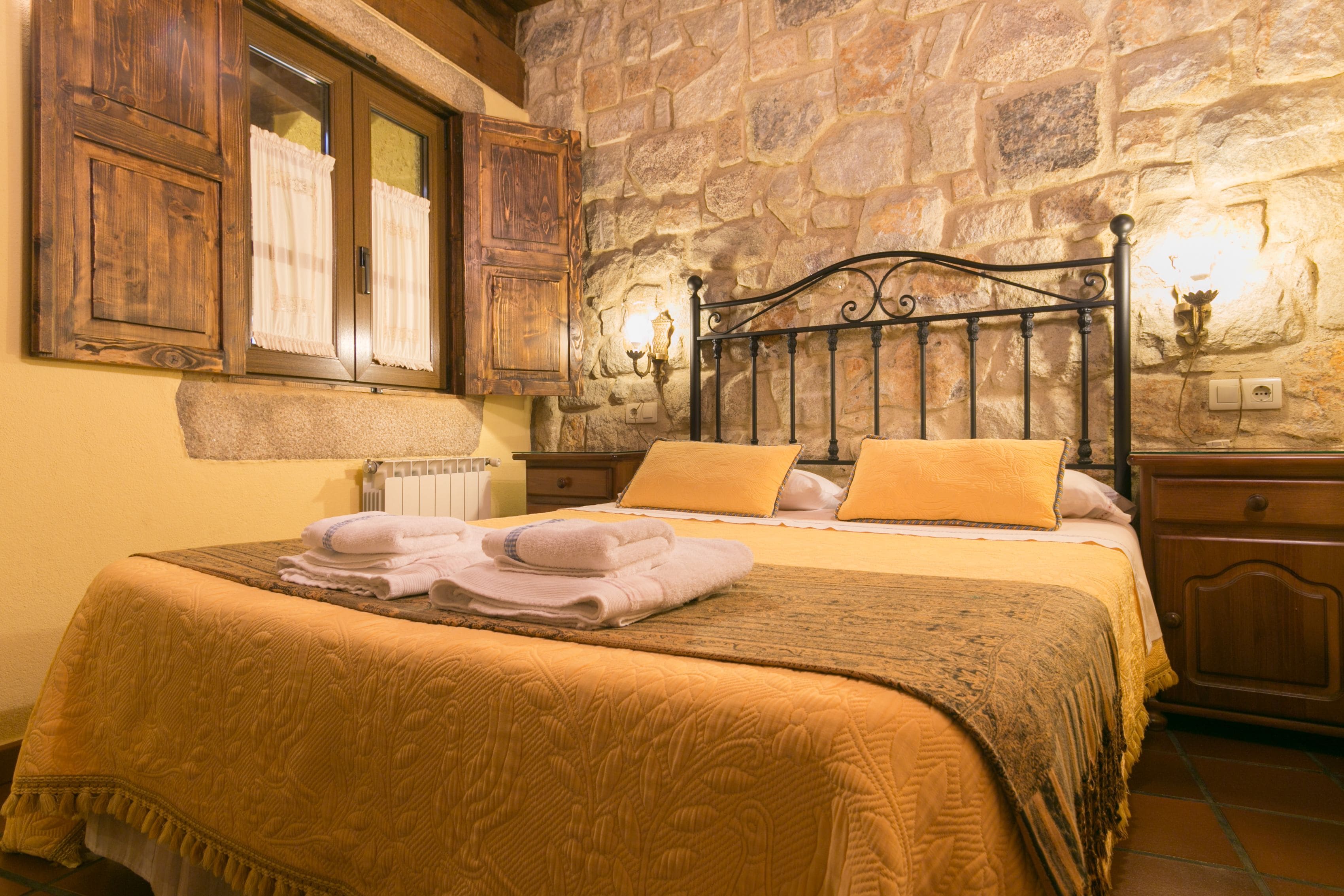 A bedroom with a stone wall, a wooden bed frame, and a window with curtains.
