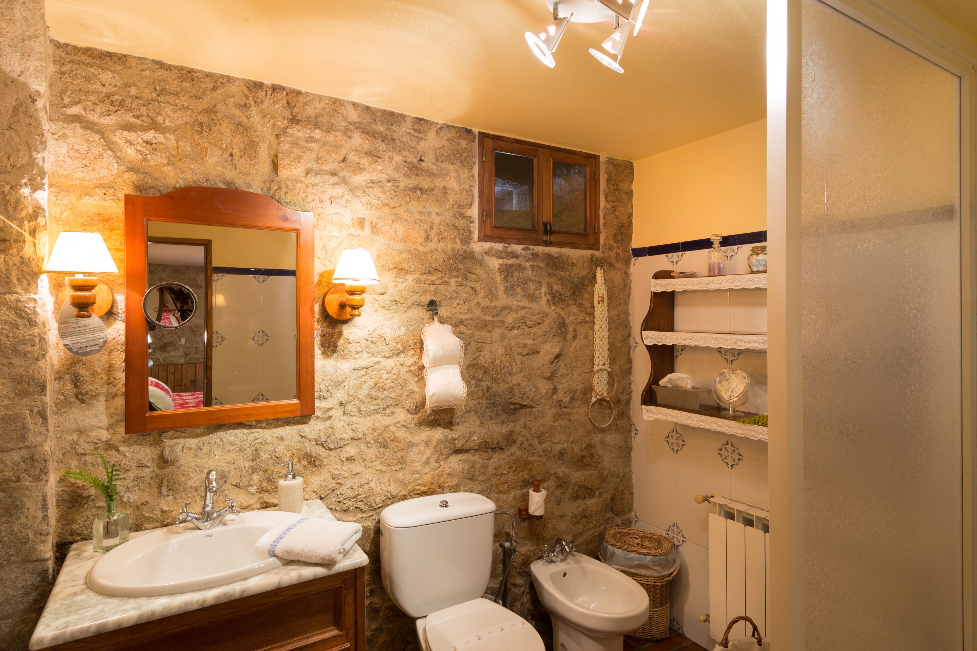Een badkamer met een stenen muur, een spiegel met houten lijst, een wastafel, een toilet en een douchetoilet.