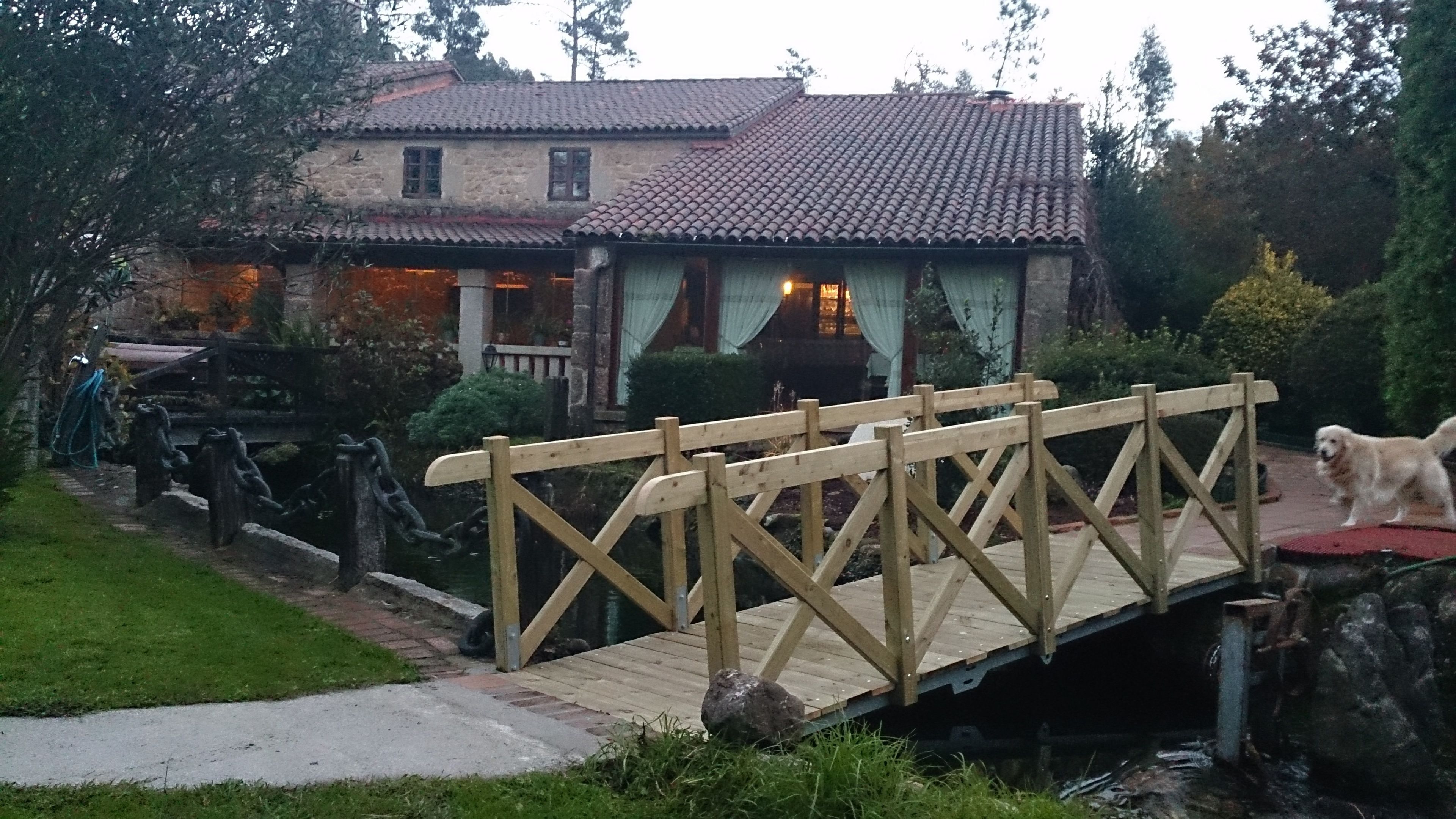 Un pont en bois enjambe un plan d’eau et mène à une maison en pierre surmontée d’un toit de tuiles.