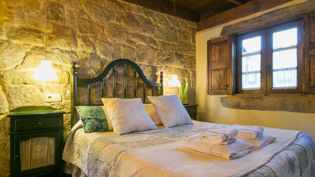 A bedroom with a stone wall, wooden headboard, and a window with curtains. There is a bedside table with a lamp and a green pillow. - Aldea Os Muiños (Rianxo)