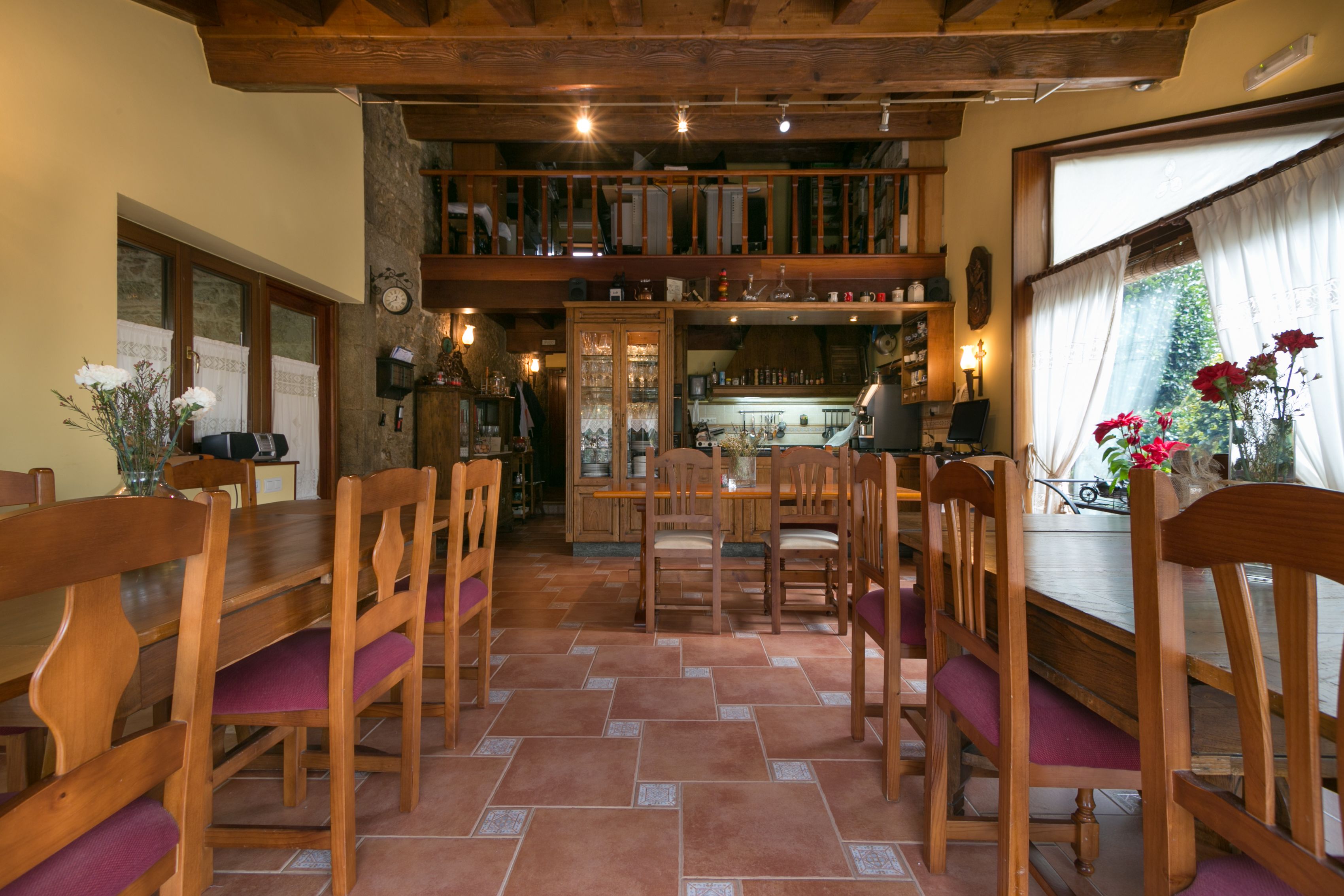 Un comedor con mesas y sillas de madera, una cocina al fondo y un ventanal con cortinas.