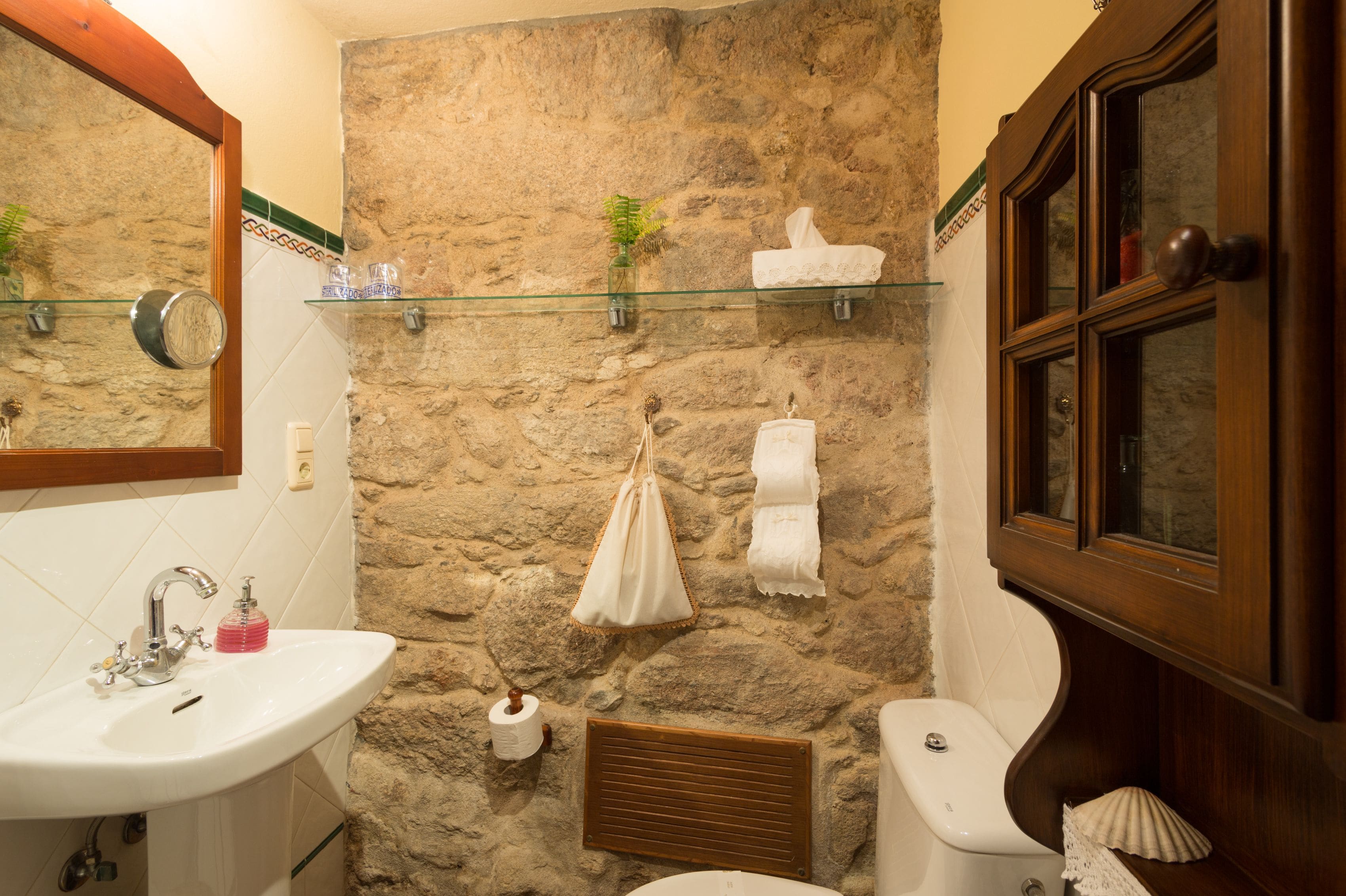 A bathroom with a stone wall, a glass shelf with toiletries, a wooden cabinet, and a sink with a mirror above it.
