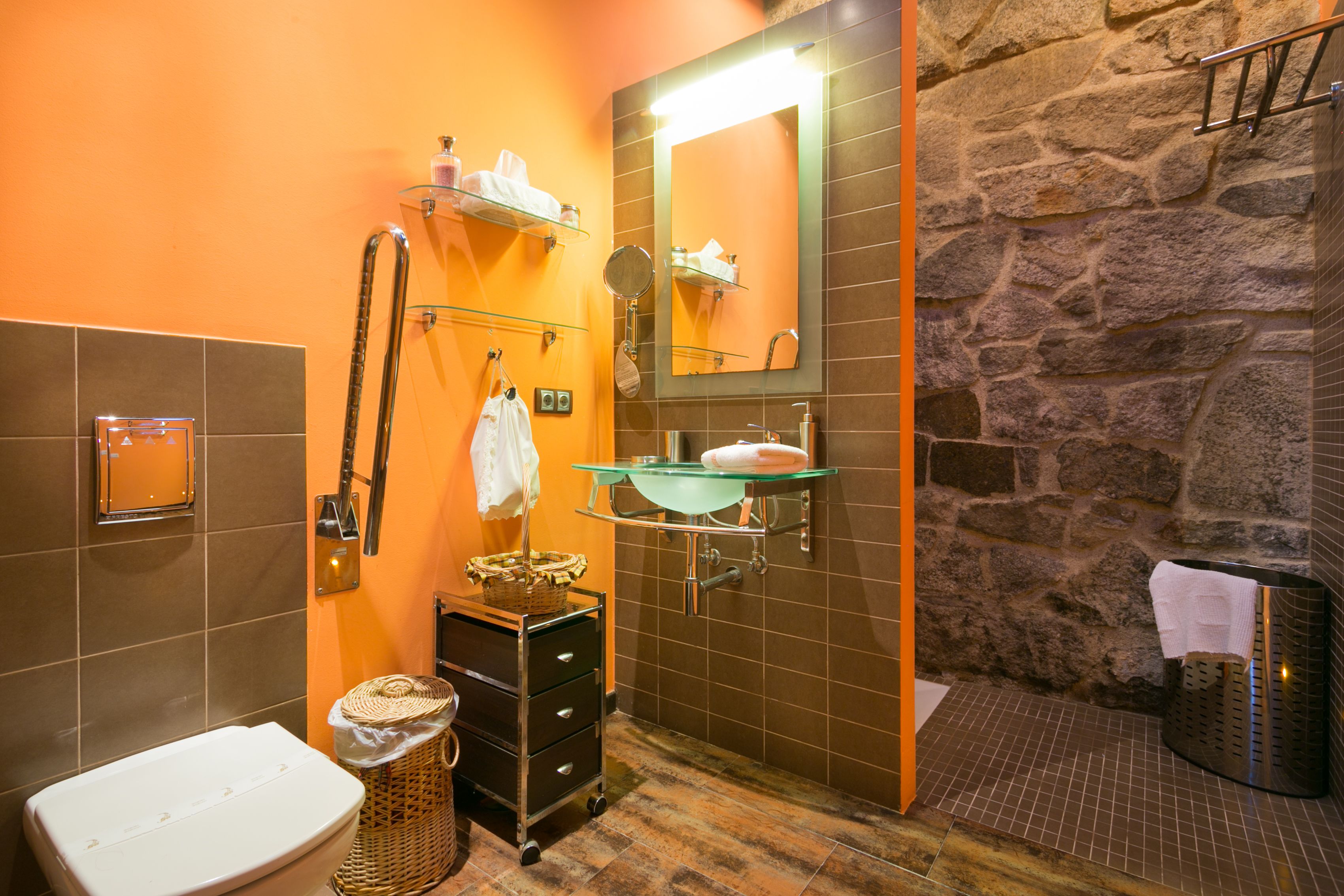 A bathroom with a stone wall, a glass sink, a mirror, a towel rack, and a toilet.