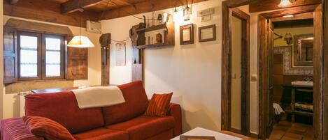 Una habitación acogedora con un sofá rojo, una mesita con un libro, vigas de madera en el techo y un baño visible a través de una puerta abierta.