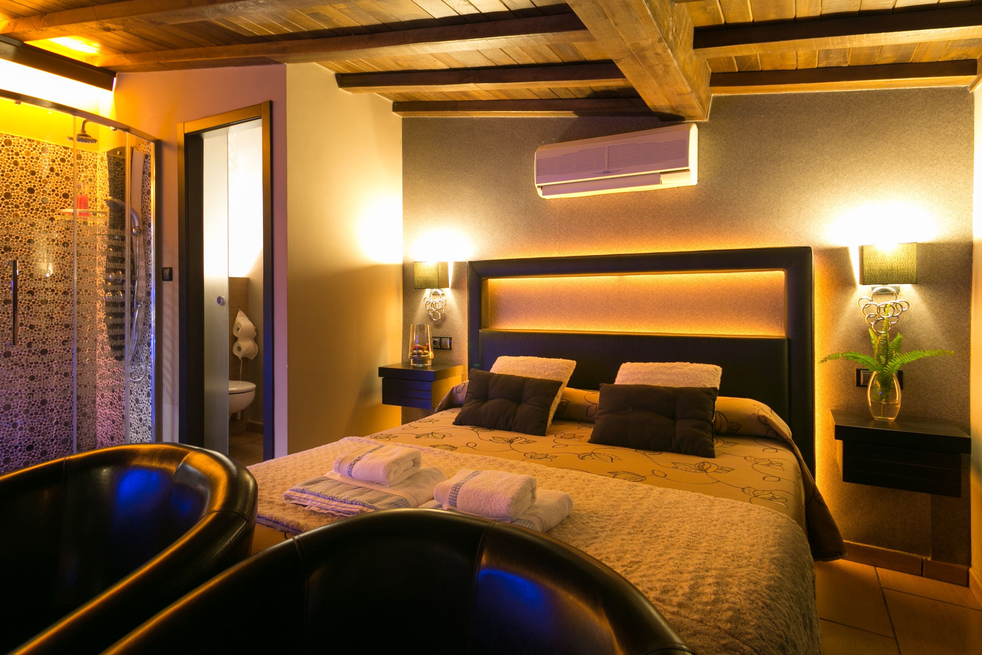 A hotel room with a large bed, a sitting area with a chair, a shower area, and a small table with a vase.