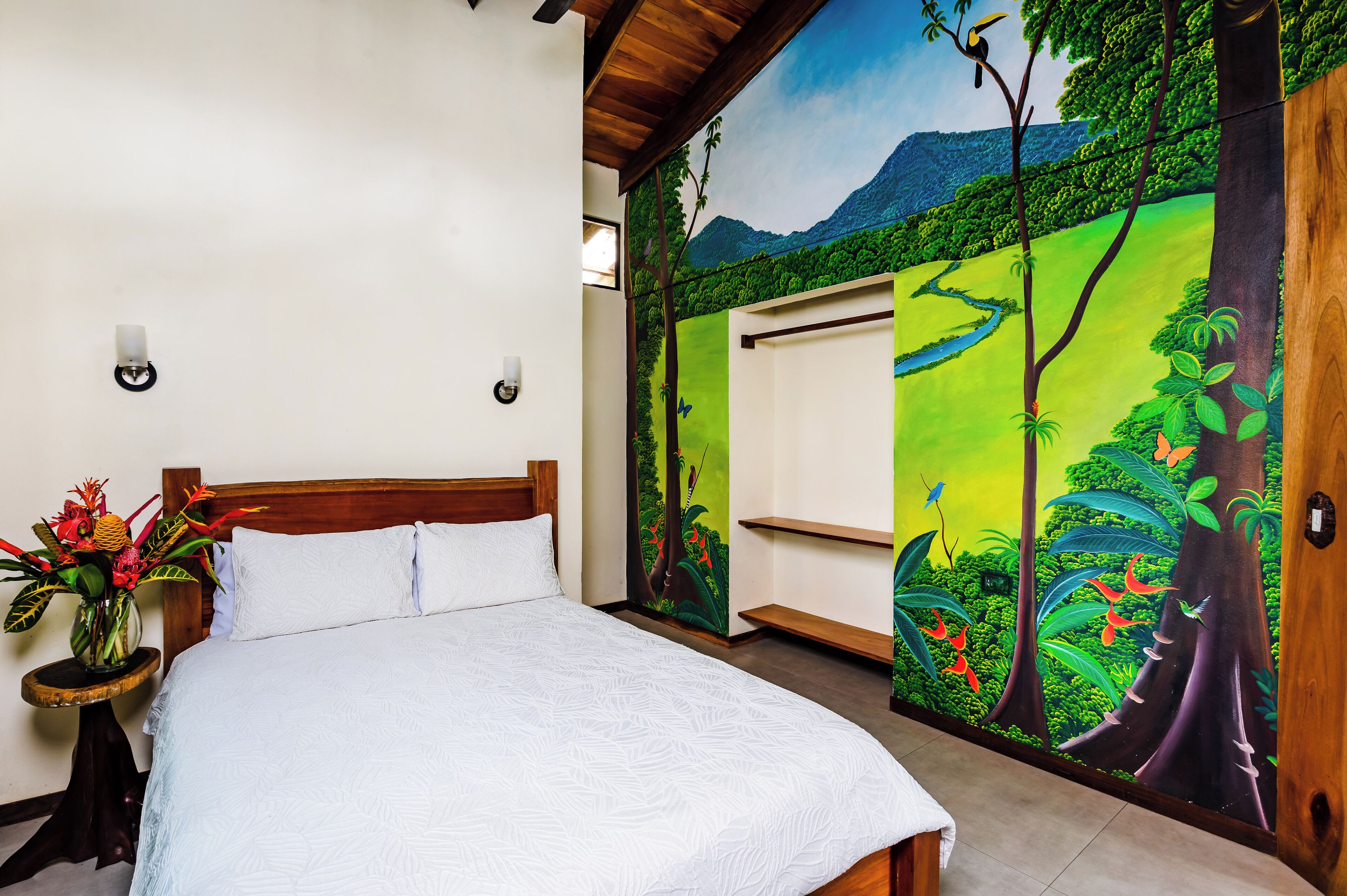 Een slaapkamer met een bed, een nachtkastje met een bloemenvaas en een muurschildering van een boslandschap.