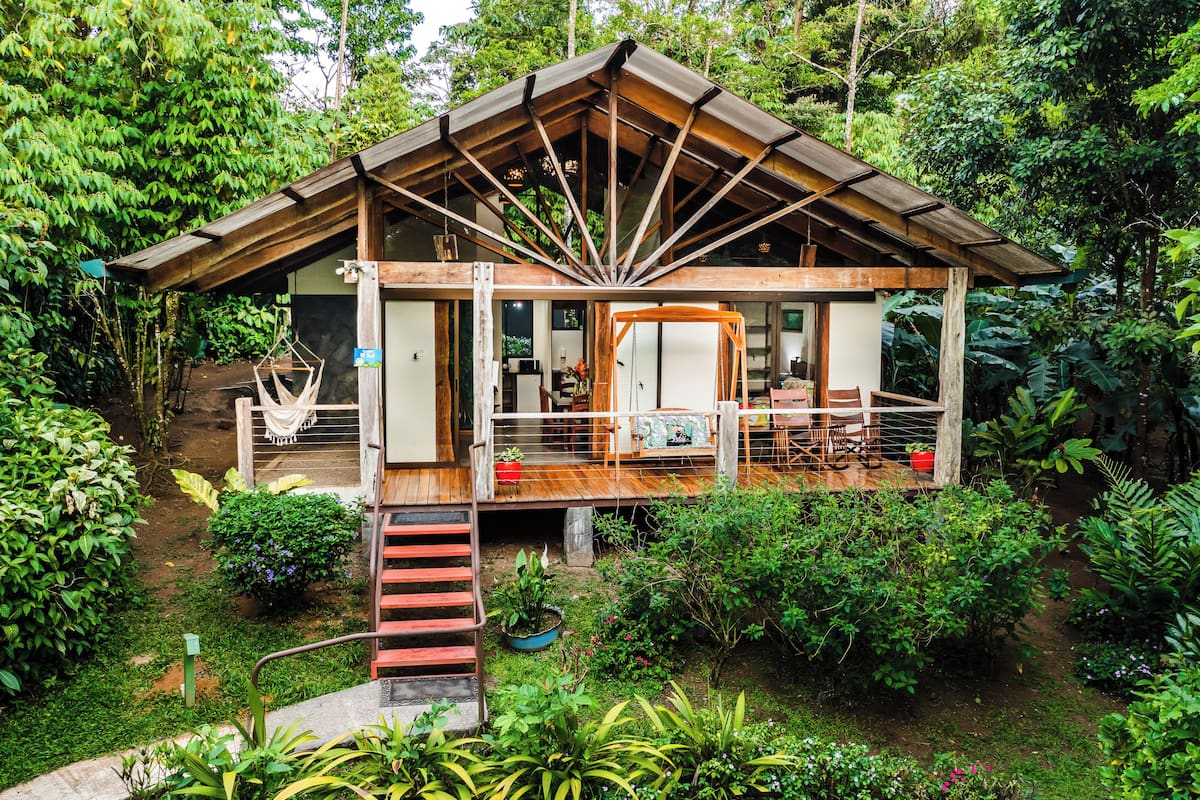 Una cabaña de madera con terraza cubierta, rodeada de abundante vegetación.