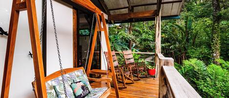 A wooden deck with a swing chair and a cushioned bench, surrounded by lush greenery.