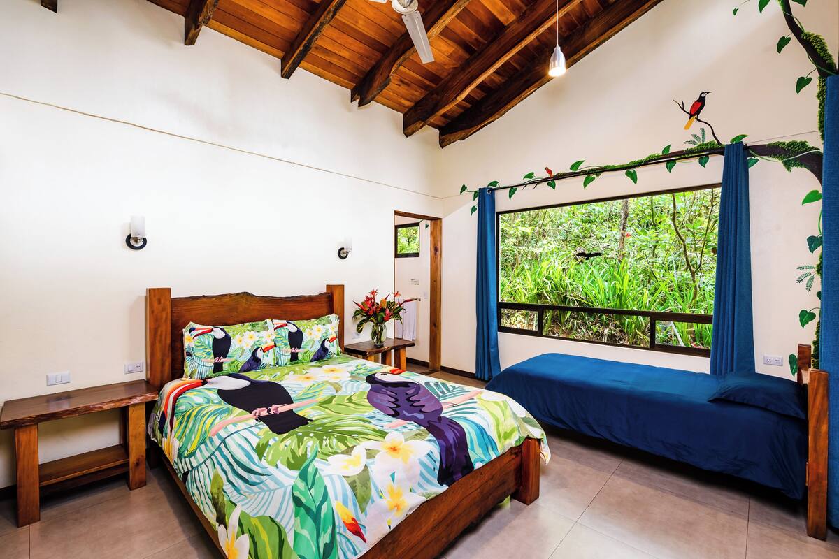 Un dormitorio con cama, un banco, una ventana con vistas a zonas verdes y un ventilador de techo.