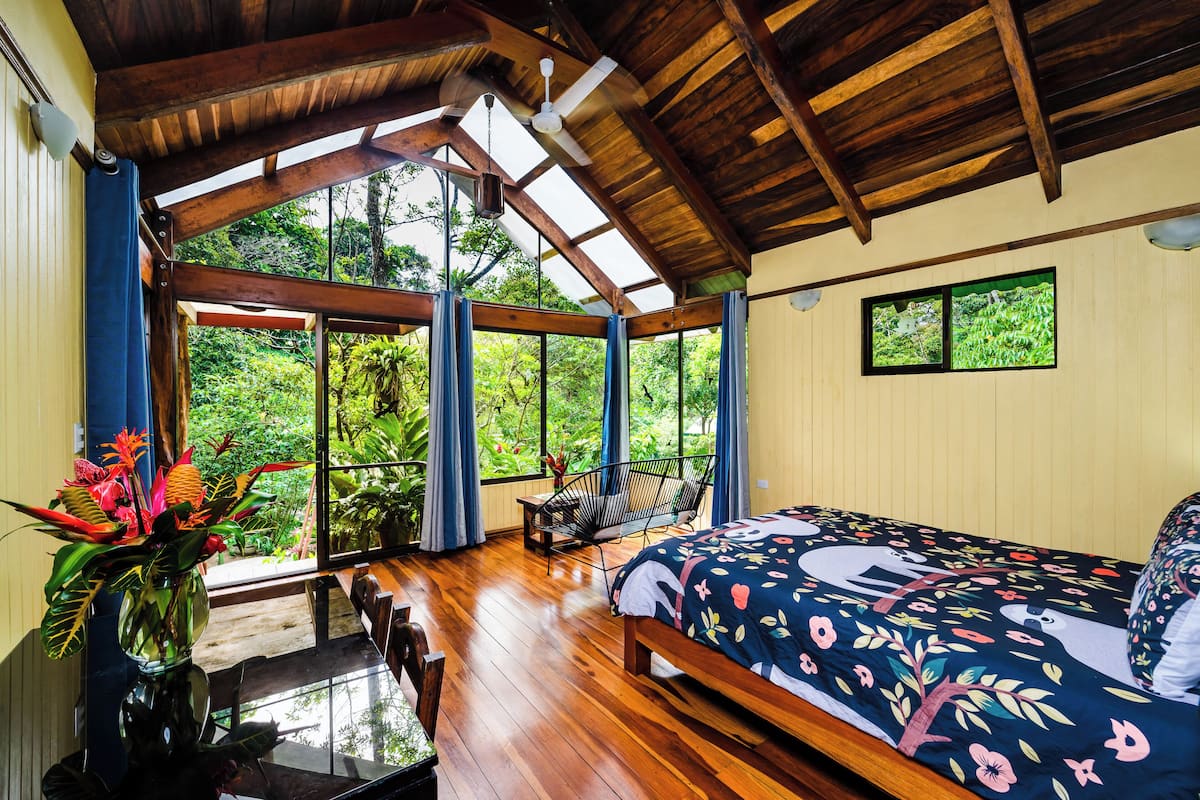 Un dormitorio con techo de madera, cama con cubrecama floral, una silla y vistas a zonas verdes a través de grandes ventanales.