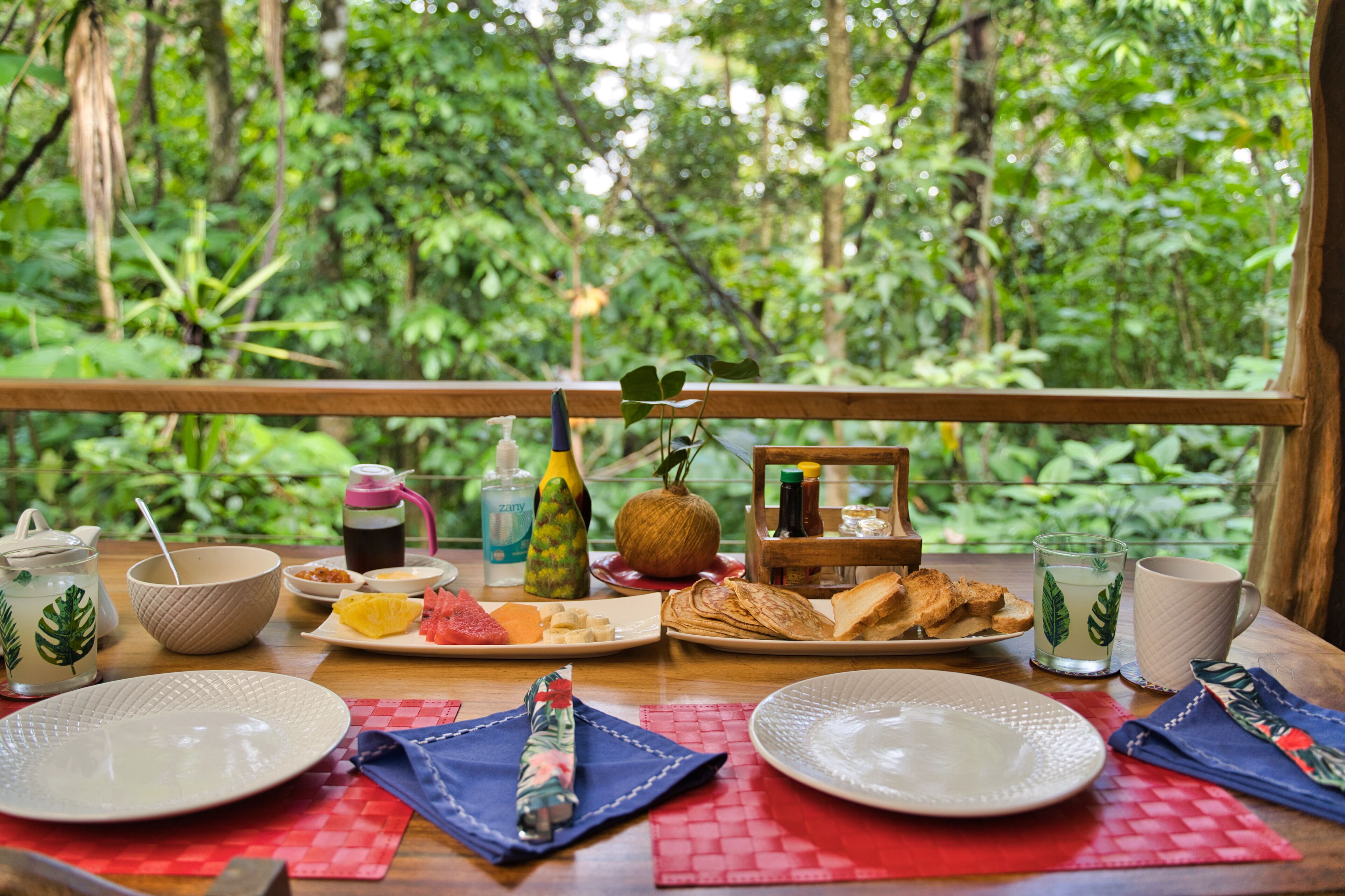 A breakfast table with various food items and drinks, set against a lush green forest backdrop.
