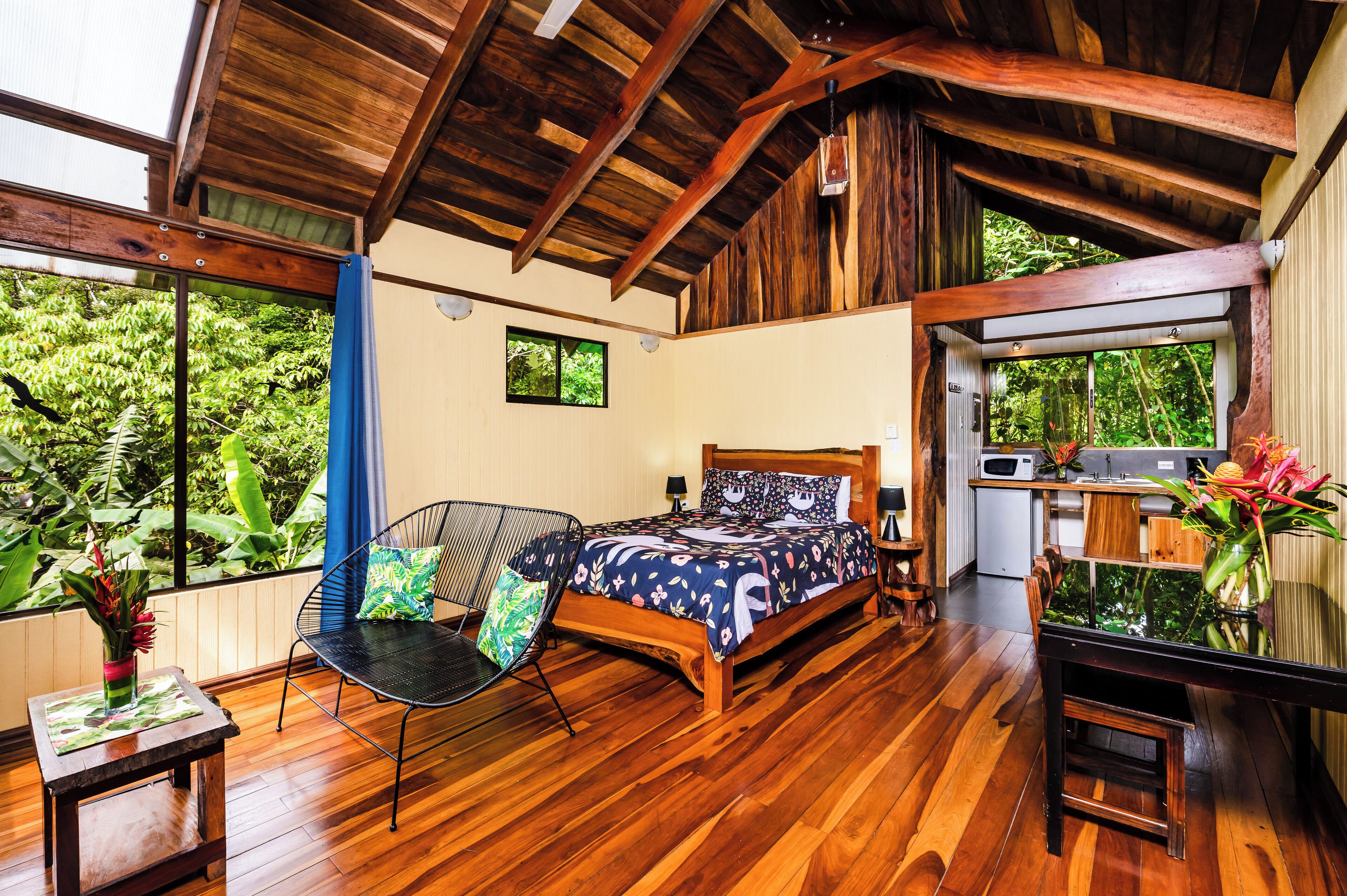 A bedroom with a wooden ceiling, a bed with floral bedding, a chair, a small table, and a view of lush greenery through large windows.