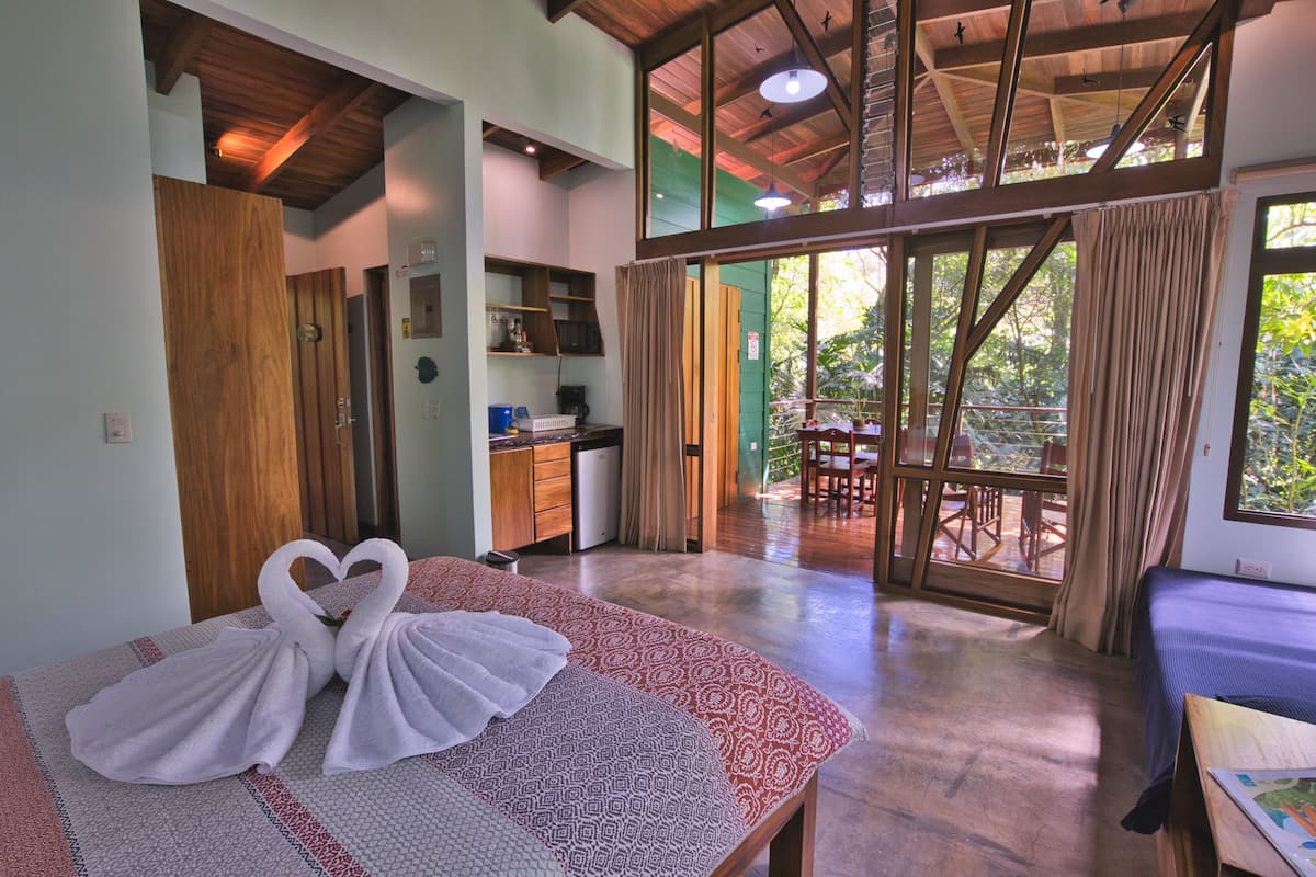 Habitación de hotel con cama, escritorio, kitchenette y balcón con mobiliario exterior.
