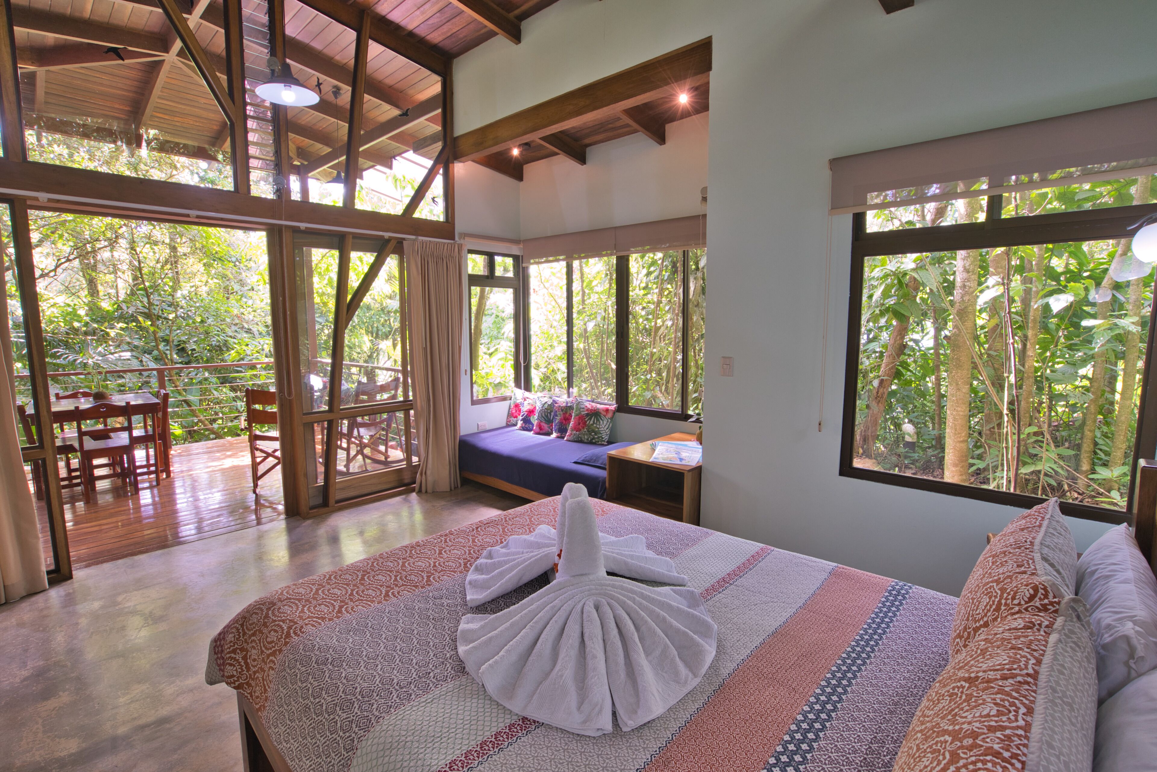 A spacious room with a large bed, a wooden ceiling, and large windows offering a view of the outdoors.