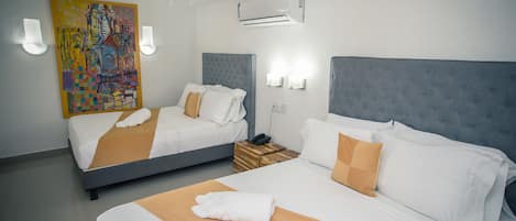 Habitación de hotel con dos camas, cada una con sábanas blancas y cabeceros marrones. Se aprecia un cuadro abstracto de colores en la pared, una mesita de noche con lámpara y un equipo de aire acondicionado sobre las camas.