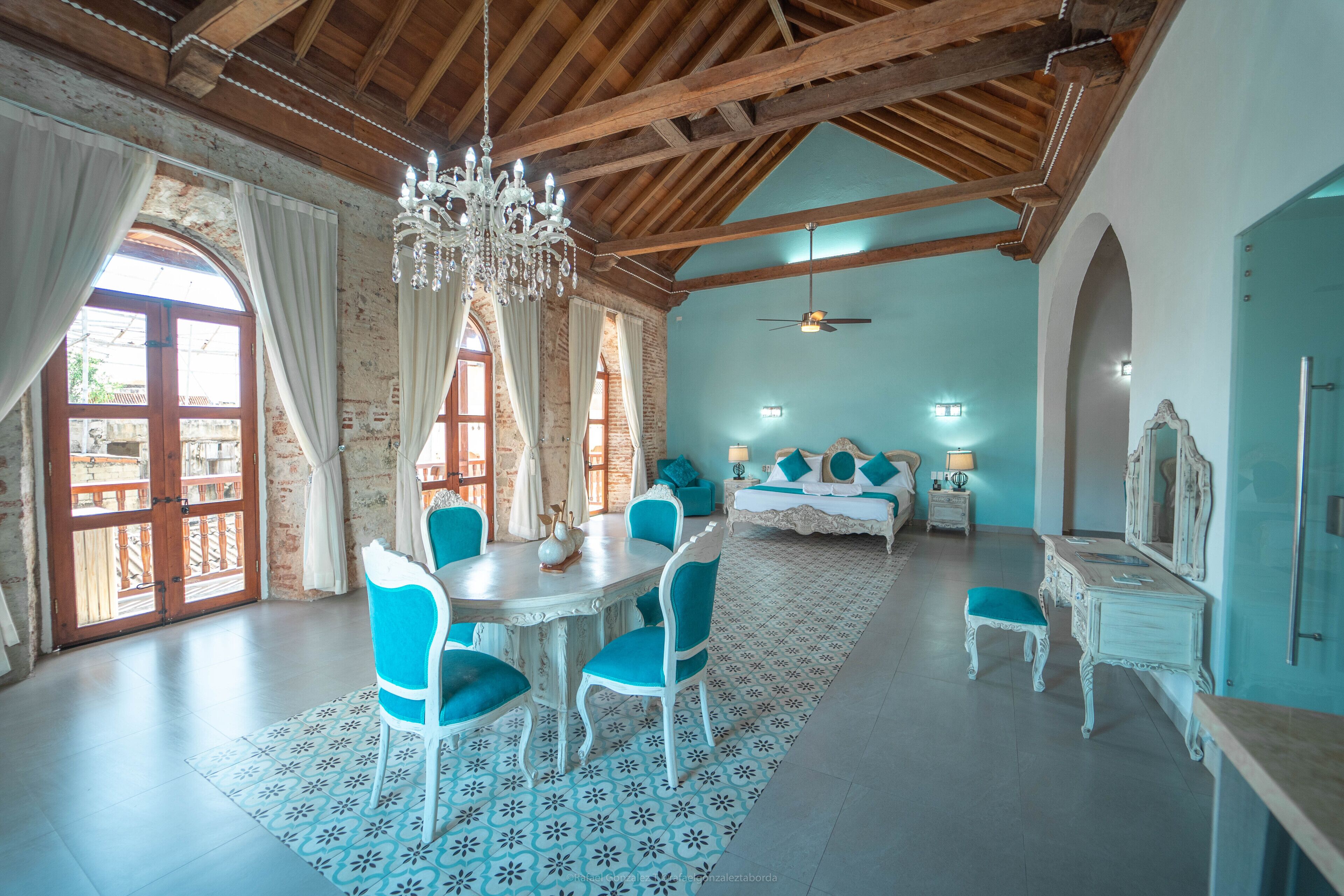 A spacious room with a high ceiling, a chandelier, a bed with a nightstand, a dresser, and a dining area with chairs and a table.