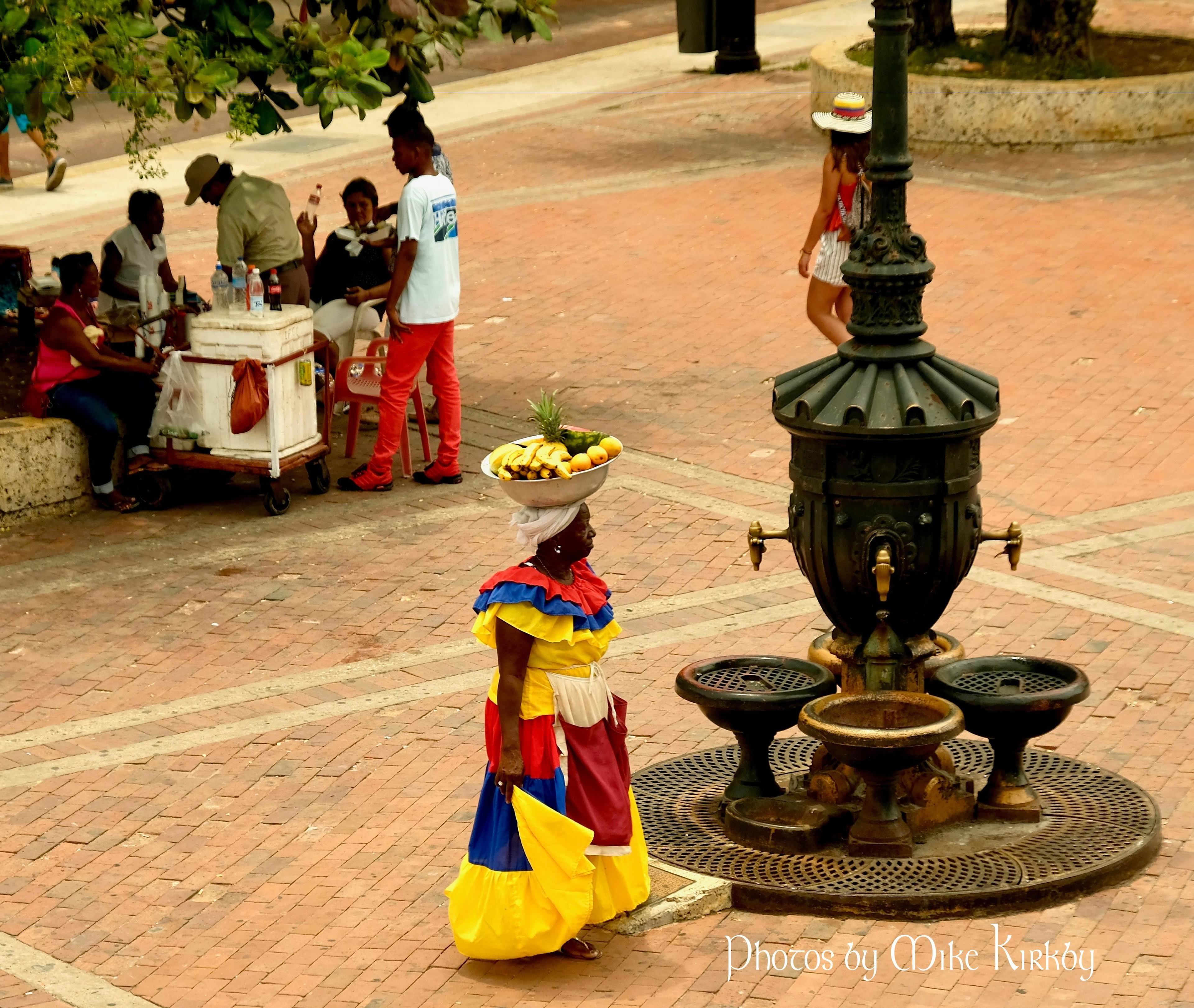 A person in a colorful dress carrying a basket of fruits, a street vendor with a cart, and a decorative fountain.