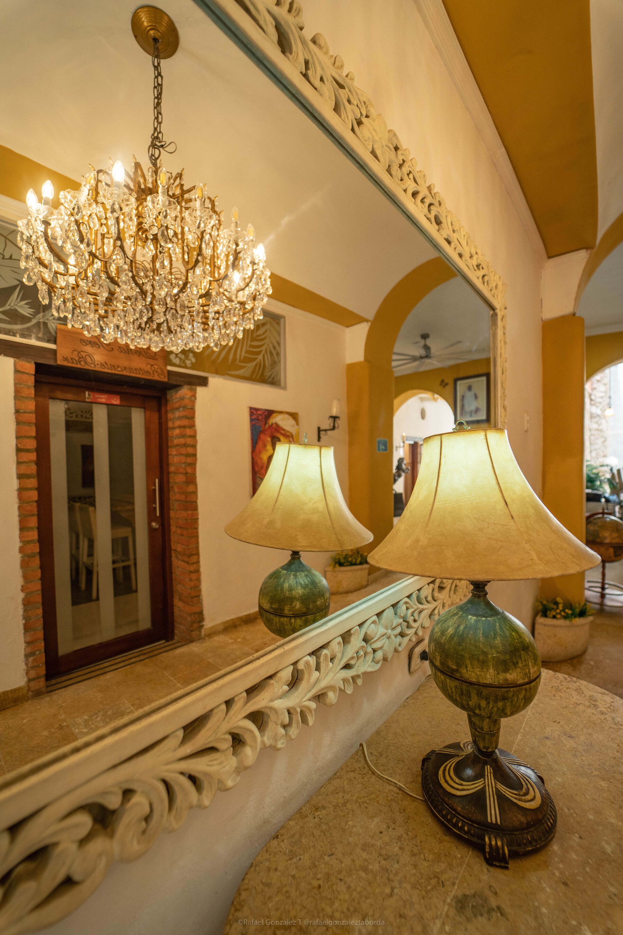 Um saguão com um candelabro, duas luminárias e um espelho refletindo o ambiente.
