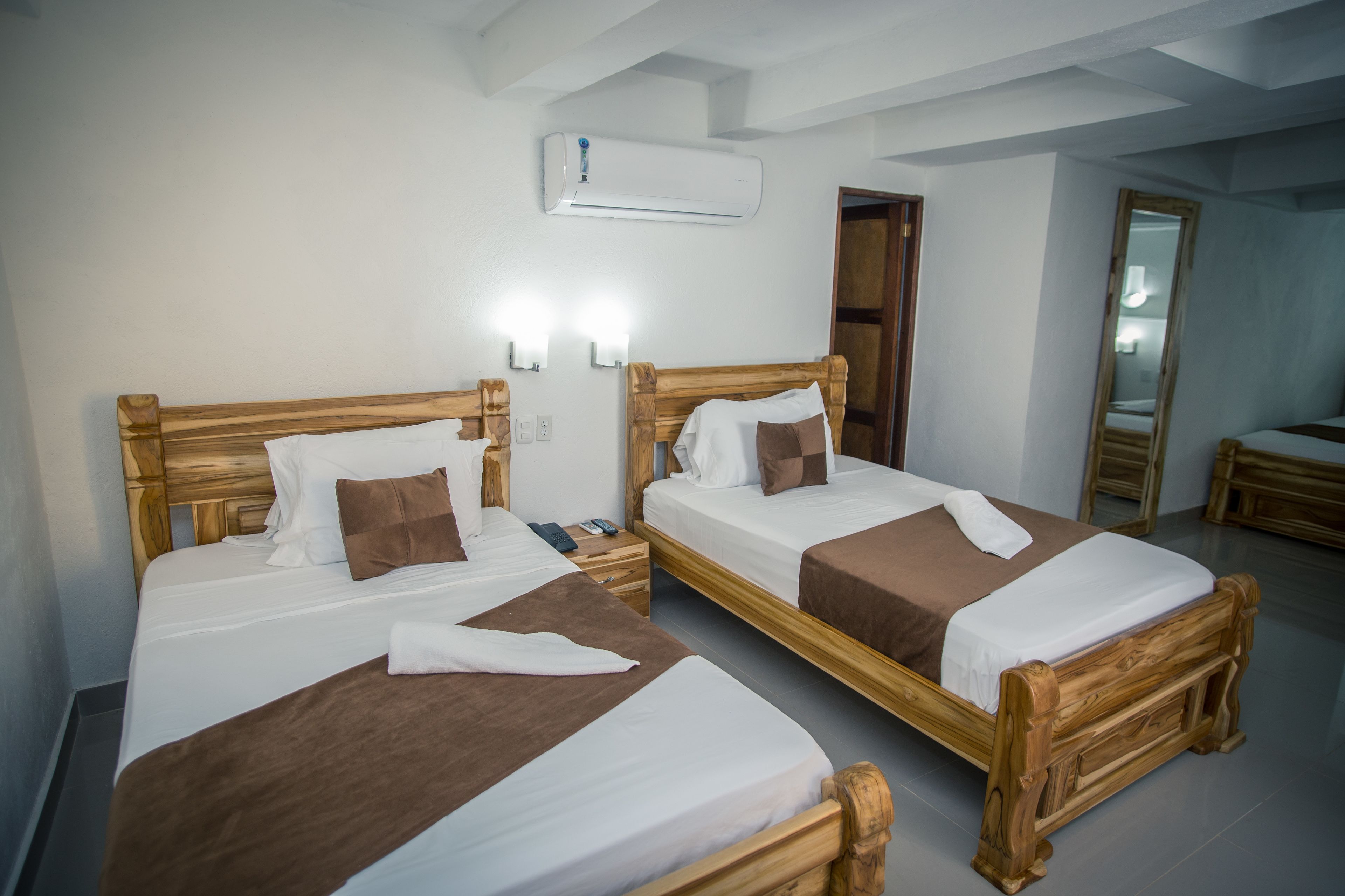 Duas camas de madeira com roupa de cama branca e castanha, uma mesa de cabeceira com telefone e comando, e um ar condicionado fixado na parede.