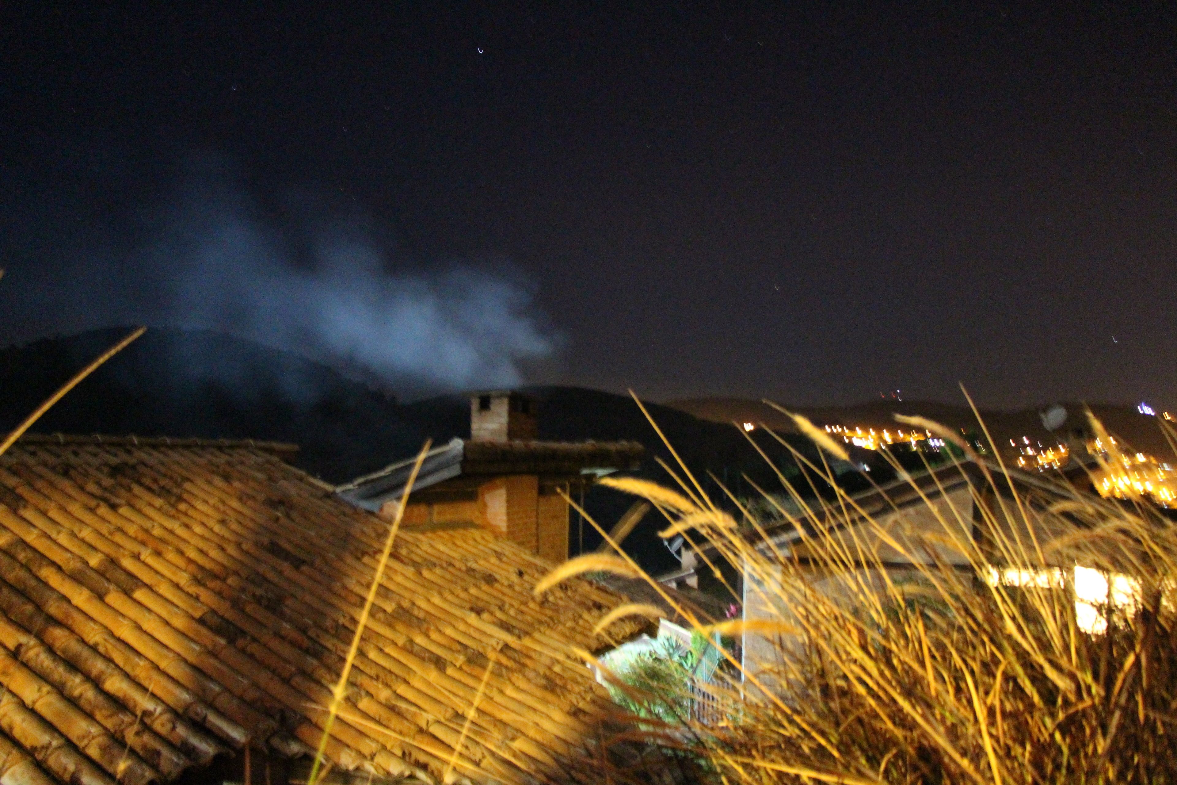 Escena nocturna con un edificio que emite humo, rodeado de hierba alta e iluminado por luces lejanas.