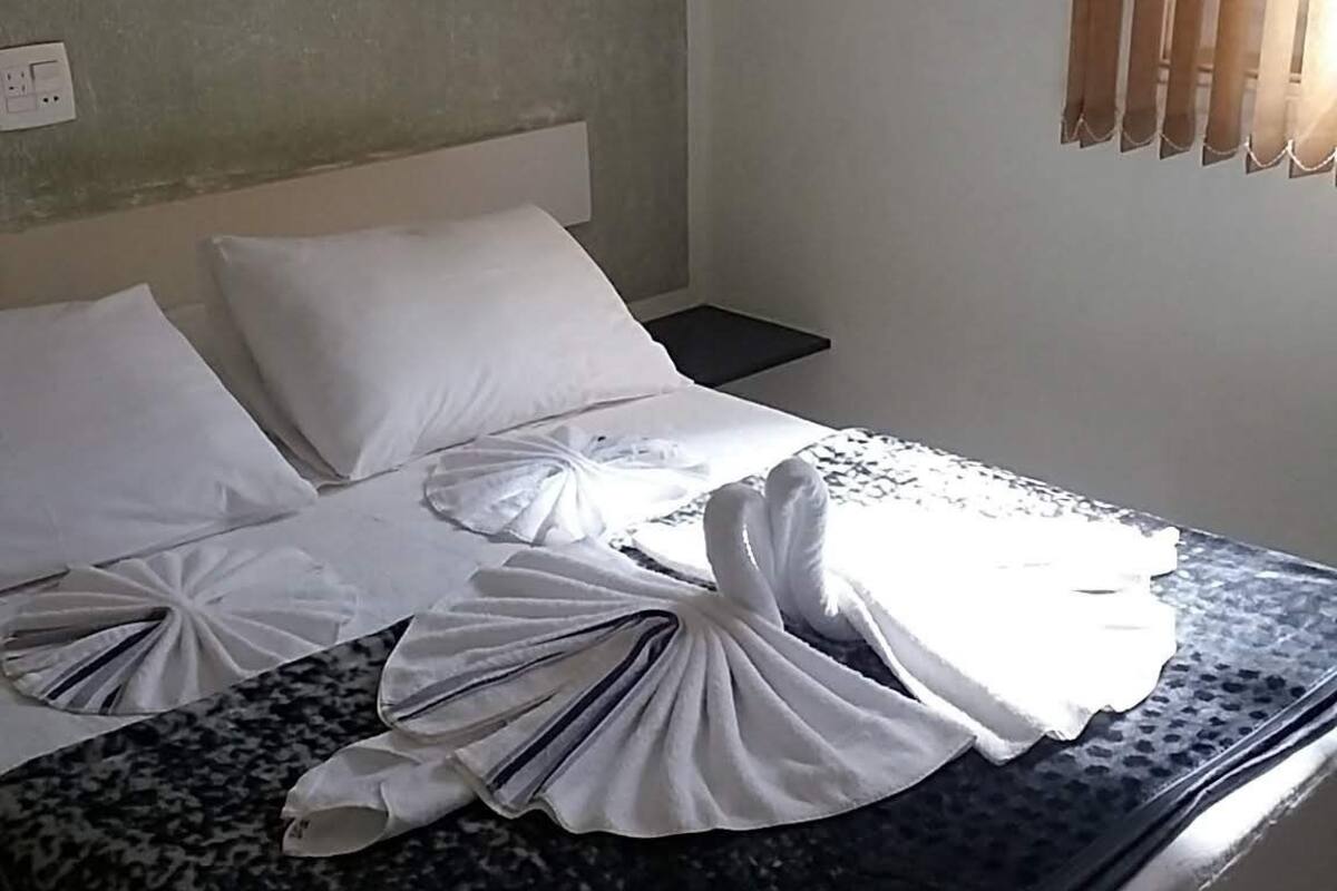 A hotel room with a bed, white pillows, a dark patterned bedspread, and a window with blinds.