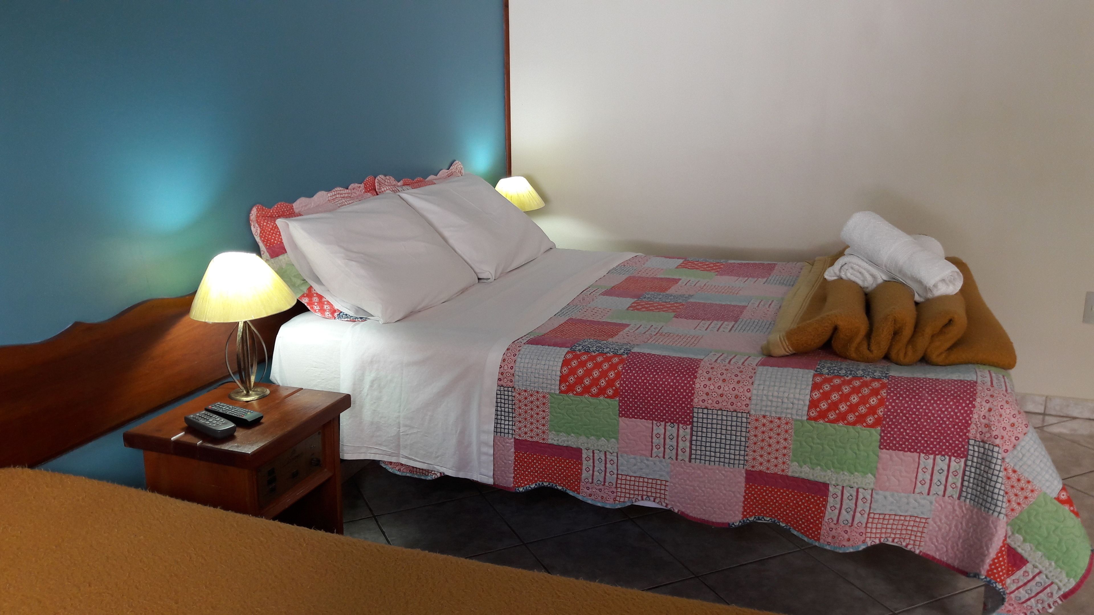 A neatly made bed with a patchwork quilt, a bedside lamp, a remote control on a wooden nightstand, and a stuffed toy on the bed.