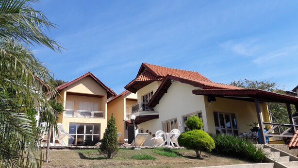 A two-story building with a red-tiled roof, a balcony, and a small garden area with chairs and a tree. - Miragem Chales (Serra Negra)